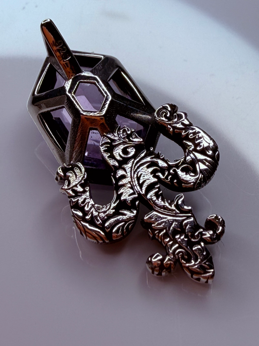An ornate metal pendant featuring a dark silver-toned geometric cage with a purple gemstone and a detailed, filigree-like silver dragon or serpent overlay, resting on a light gray surface.