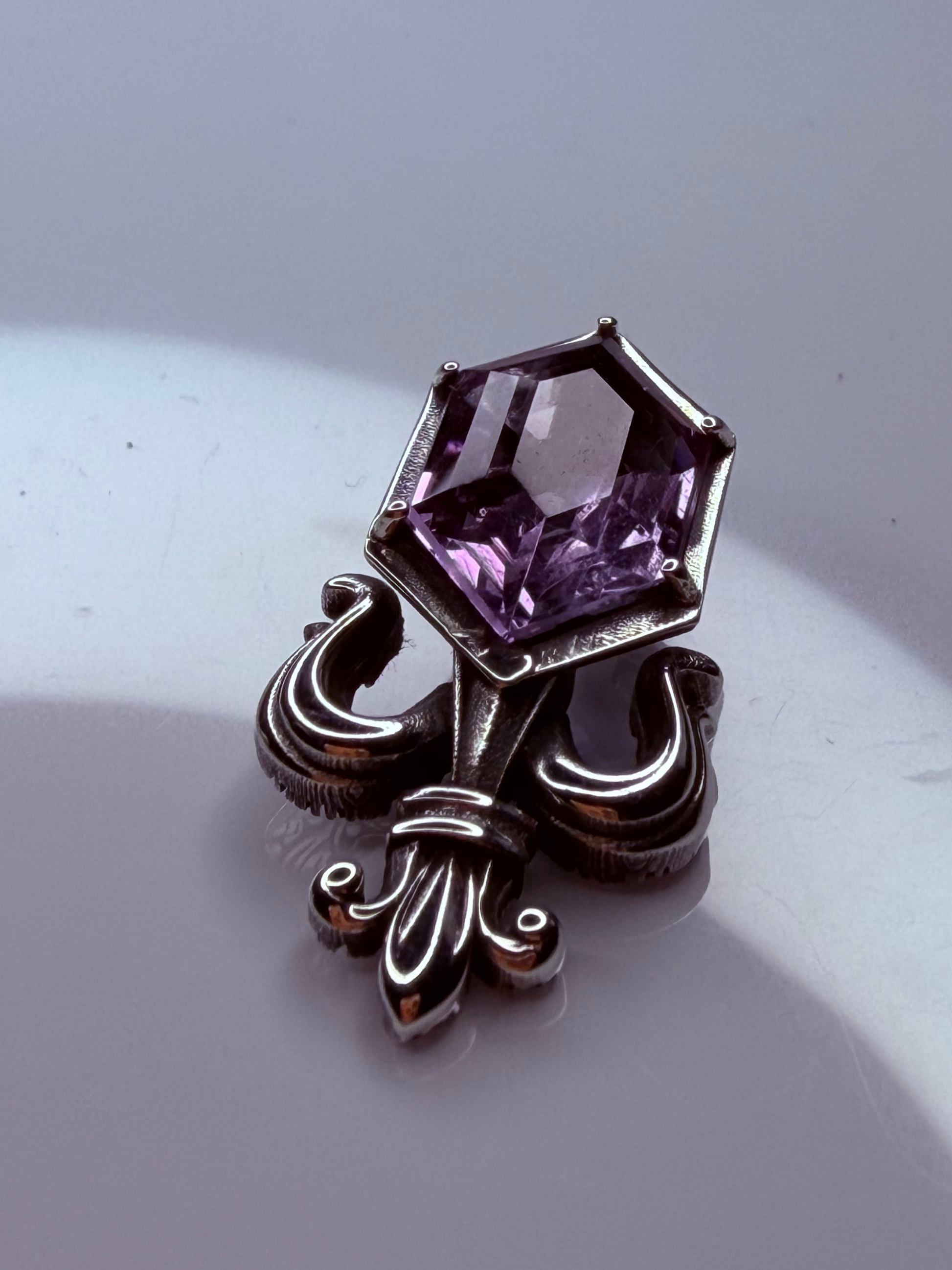 A vintage-style silver-toned brooch featuring a faceted purple gemstone, displayed on a light gray surface with a soft shadow.
