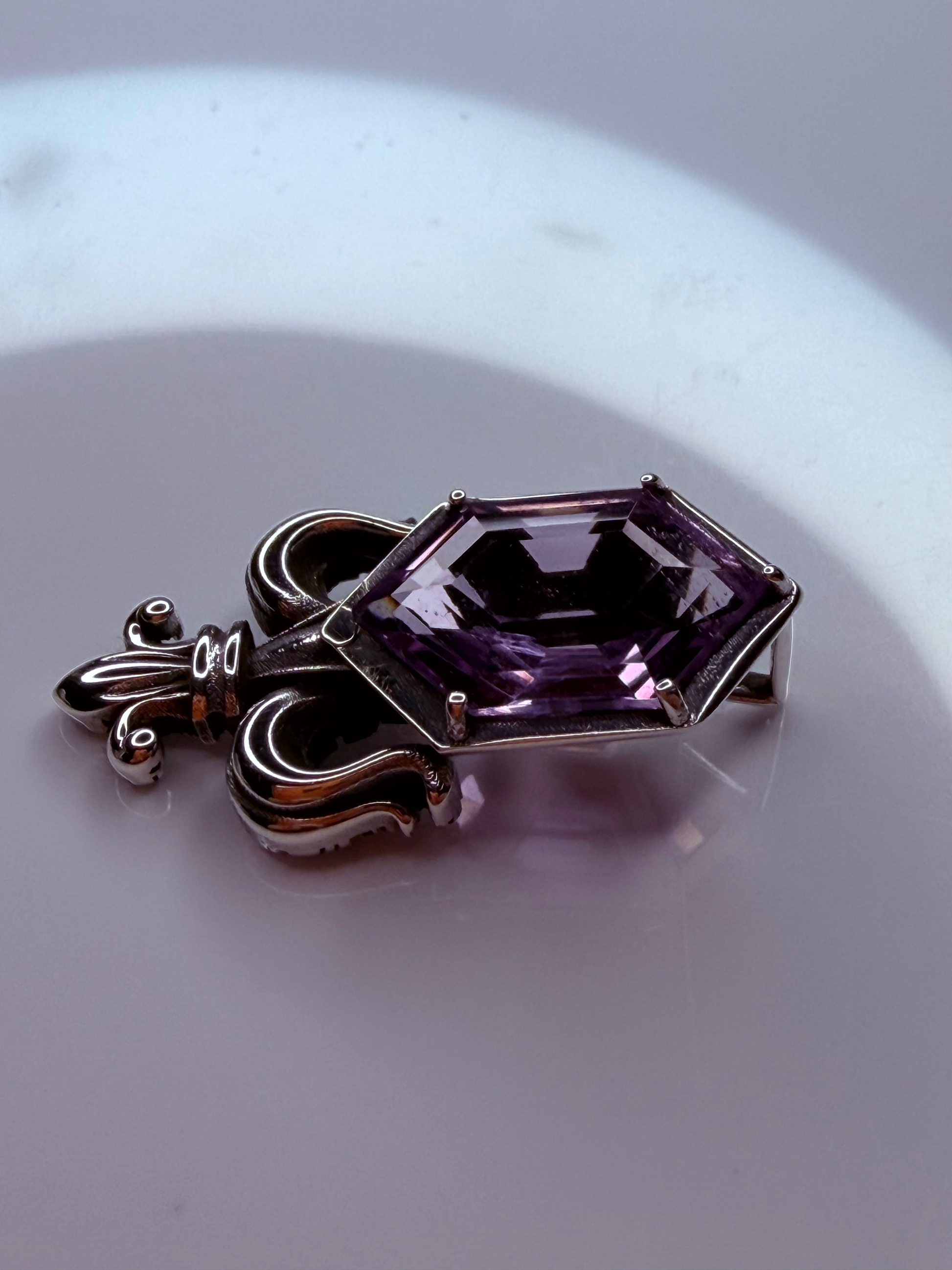 A silver-toned pendant featuring a faceted purple amethyst gemstone, resting on a reflective light gray surface.