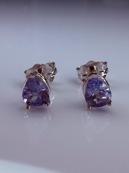 A pair of pear stud earrings featuring faceted tanzanite gemstones in a white gold setting, displayed on a reflective white surfce. 